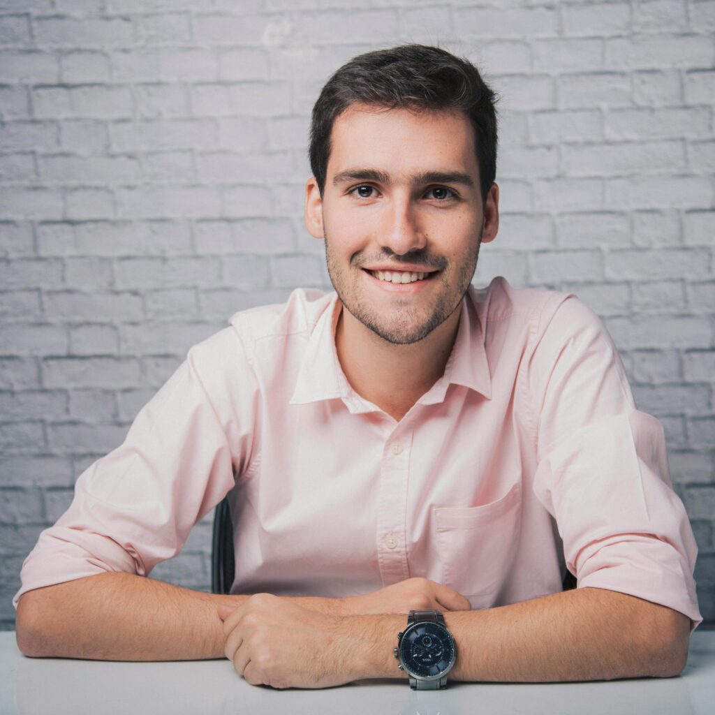 Positive guy crossing arms while sitting on chair against white brick wall and looking at camera