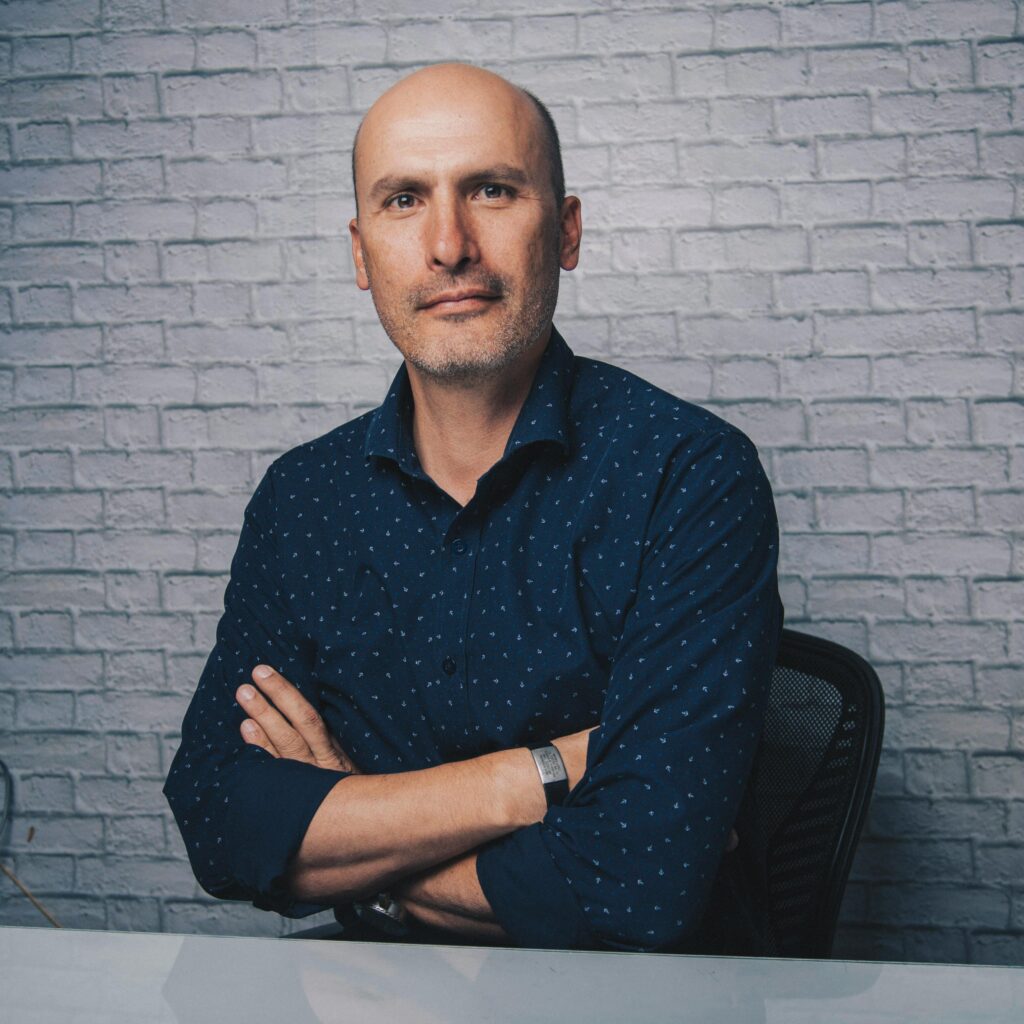 Positive bald male in blue shirt sitting at shiny desk with crossed arms while looking at camera in white room